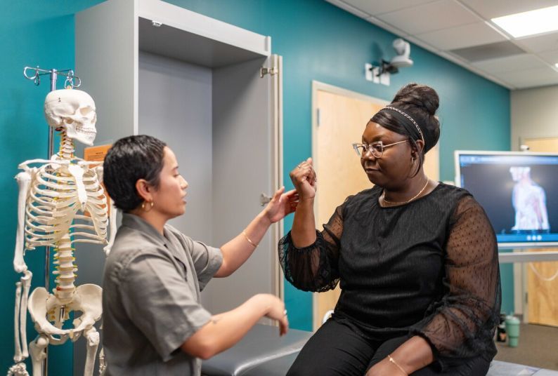 One student sits on a bench while another student examines her arm. 